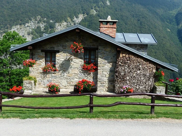 A stone house with a stacked woodpile and flowers in a mountainous setting.